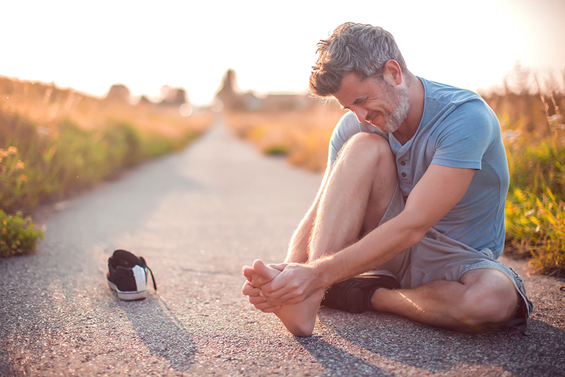 man-holding-foot-farm-country Man experiencing foot pain while sitting on a walking path and holding his injured foot.